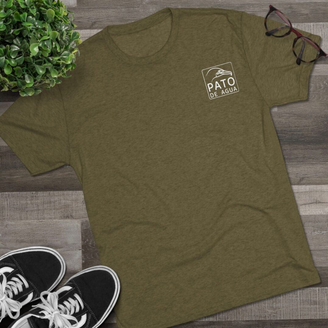Olive green t-shirt with a small white logo on the chest, laid on a wooden floor.