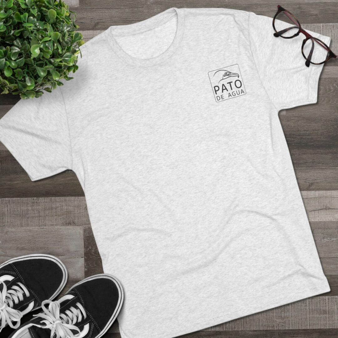 White t-shirt with a small logo, black shoes, glasses, and a plant on a wooden floor.